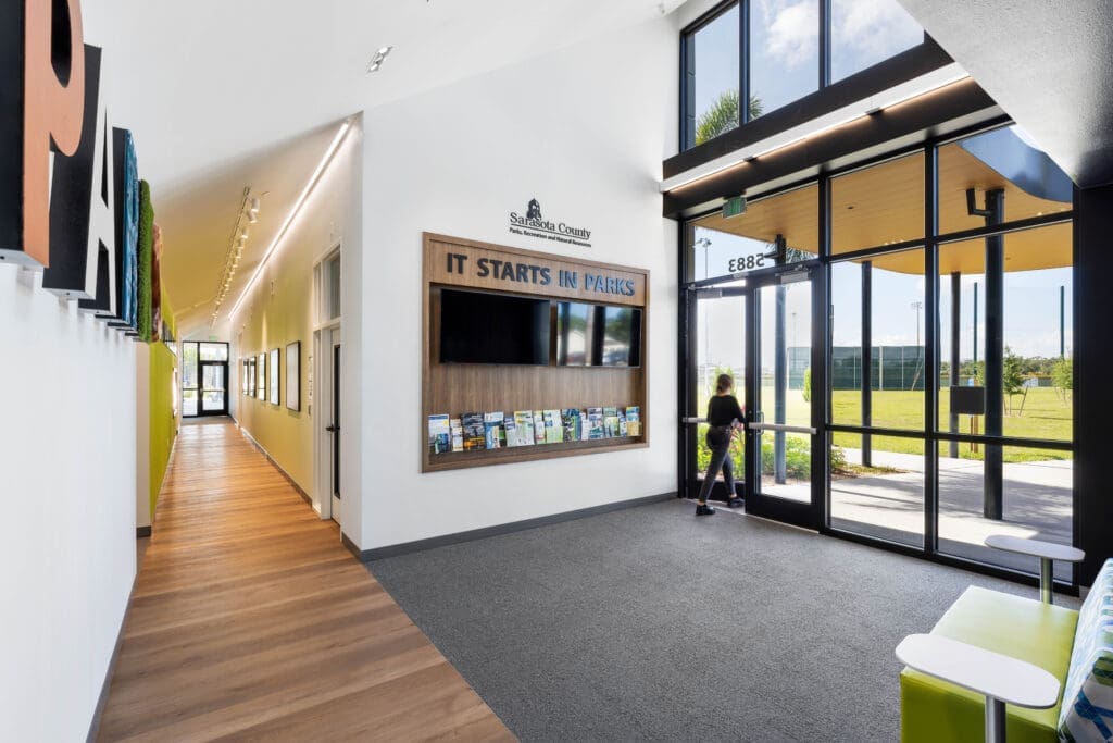 Lobby of the Sarasota County Parks Administration Building with an "It Starts in Parks" display wall, lime-green accent corridor, and floor-to-ceiling entrance glazing.