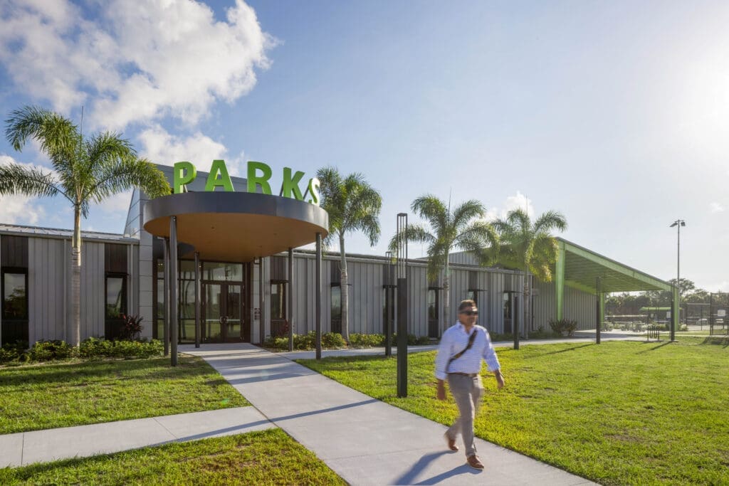 Front entrance of the Sarasota County Parks Administration Building with green "PARKS" rooftop lettering, a round metal canopy, and palm-lined walkway.