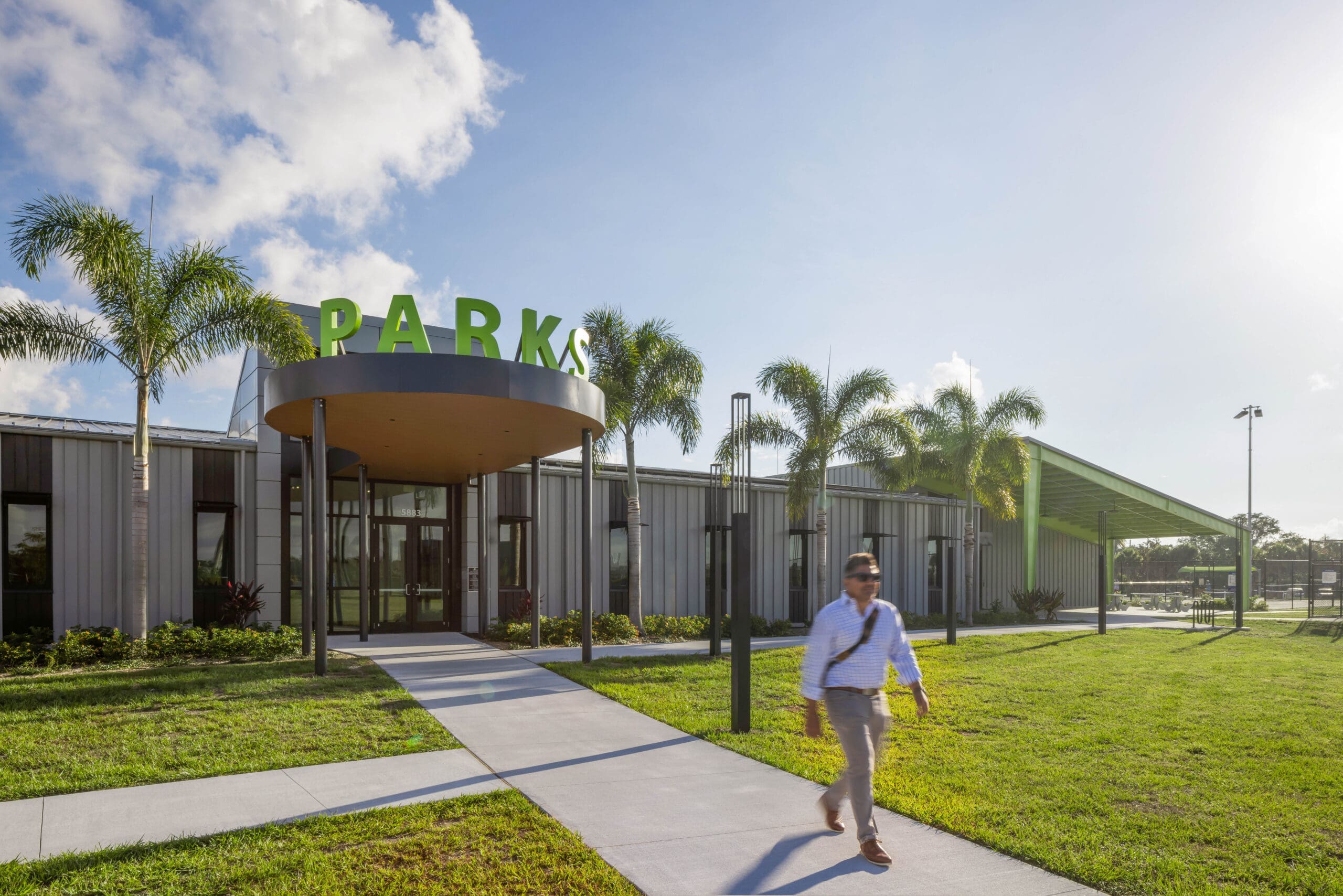 Front entrance of the Sarasota County Parks Administration Building with green "PARKS" rooftop lettering, a round metal canopy, and palm-lined walkway.